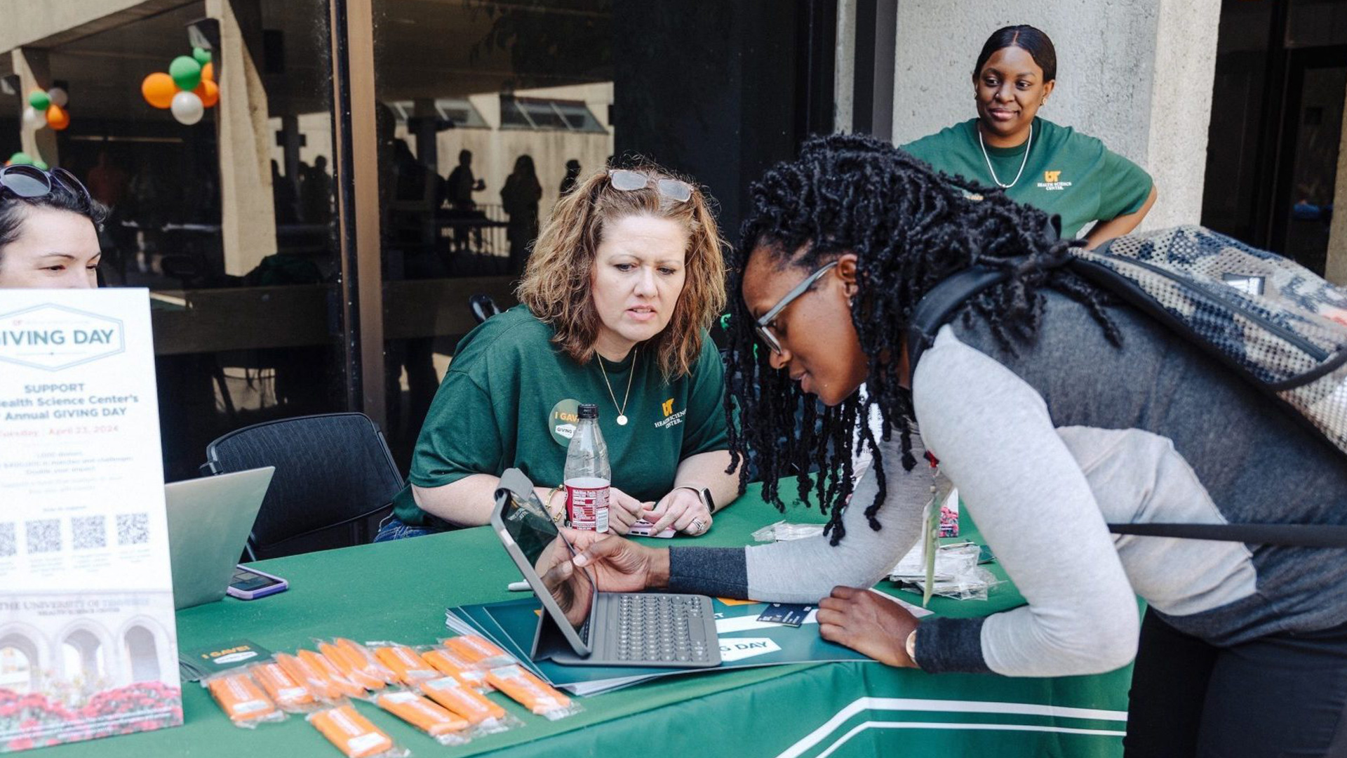 A black female student using an iPad at a table manned by women wearing green shirts.