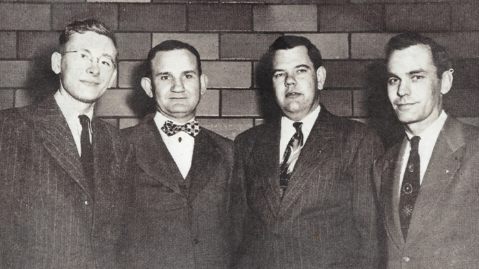 A vintage, black and white photo, showing four white men wearing suits, standing side-by-side.