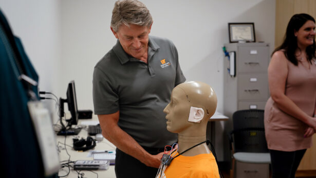 A man wearing a gray polo observes a test dummy used to aid audiology and speech pathology research.