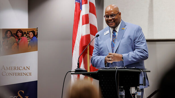 A black male wearing a sky blue suit speaks from behind a podium.