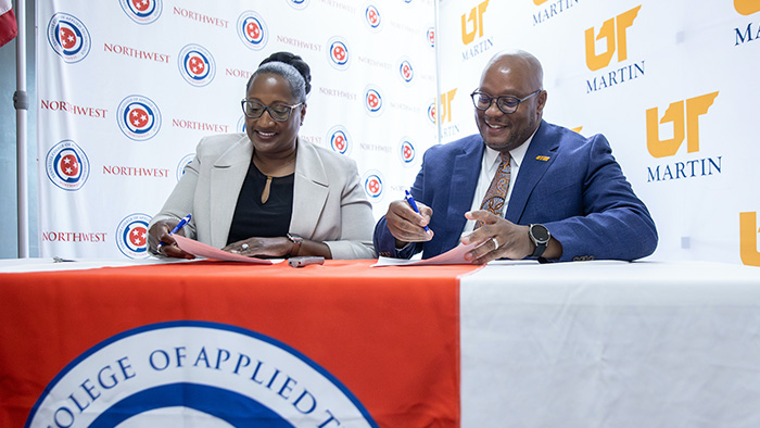 A black man and woman sit at a table signing documents.