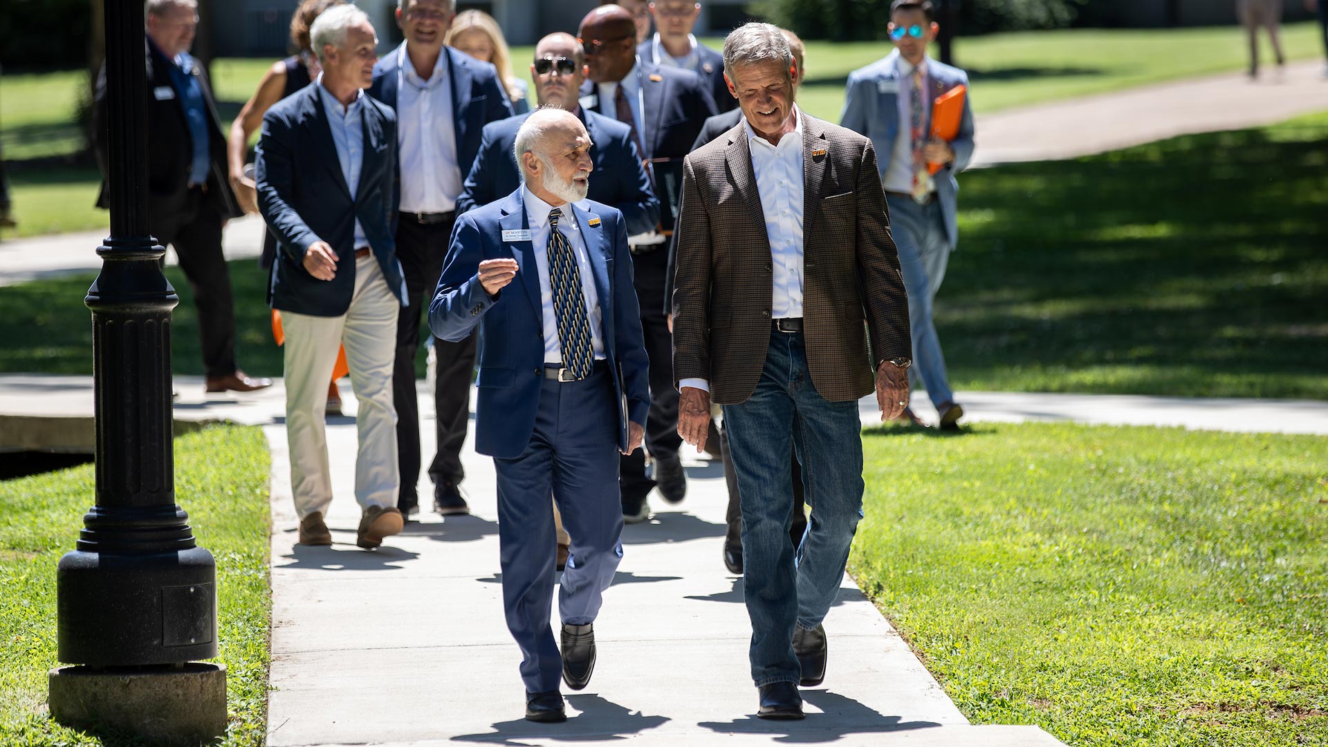 A crowd of people walk down a side walk surrounded by grass.