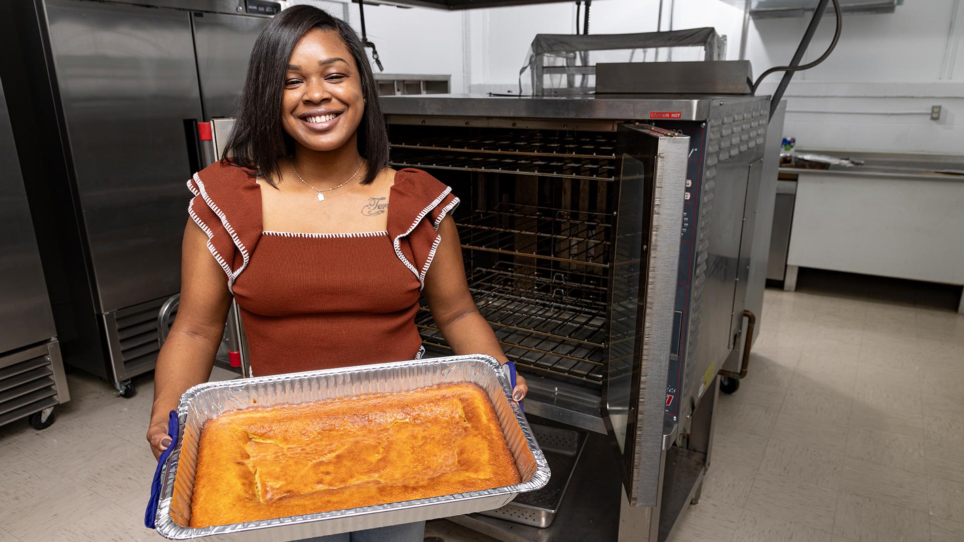 A black woman stands in a commercial kitchen, holding a pan of vegetarian cornbread.