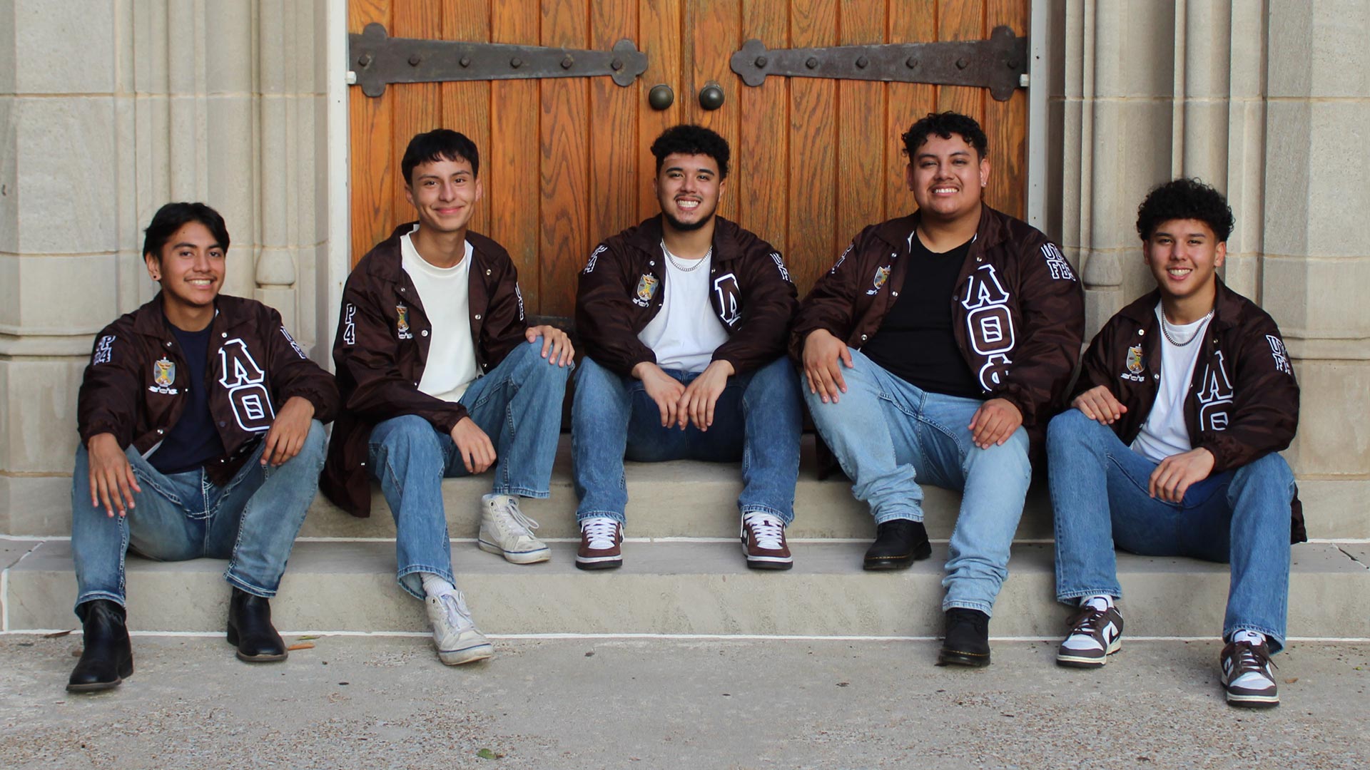 A group of hispanic young men, wearing Lambda Theta Phi jackets, sitting on steps.