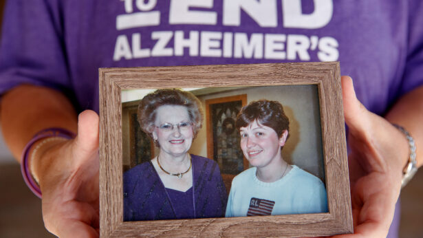 A person holds a framed photo of a young, red-haired woman standing with an elderly woman in a purple suit.