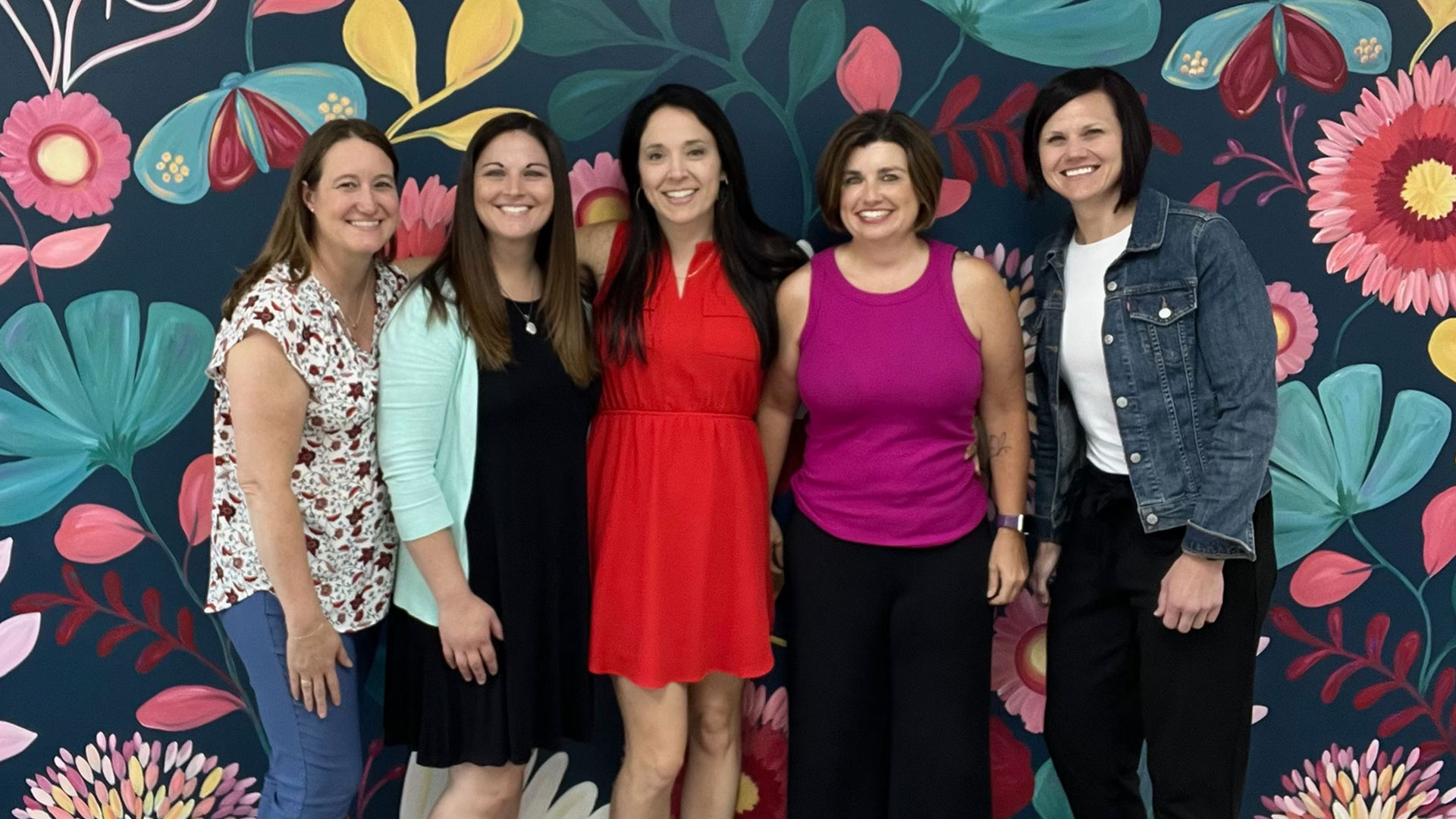 Five women, all smiling, stand in front of a wall painted with flowers.