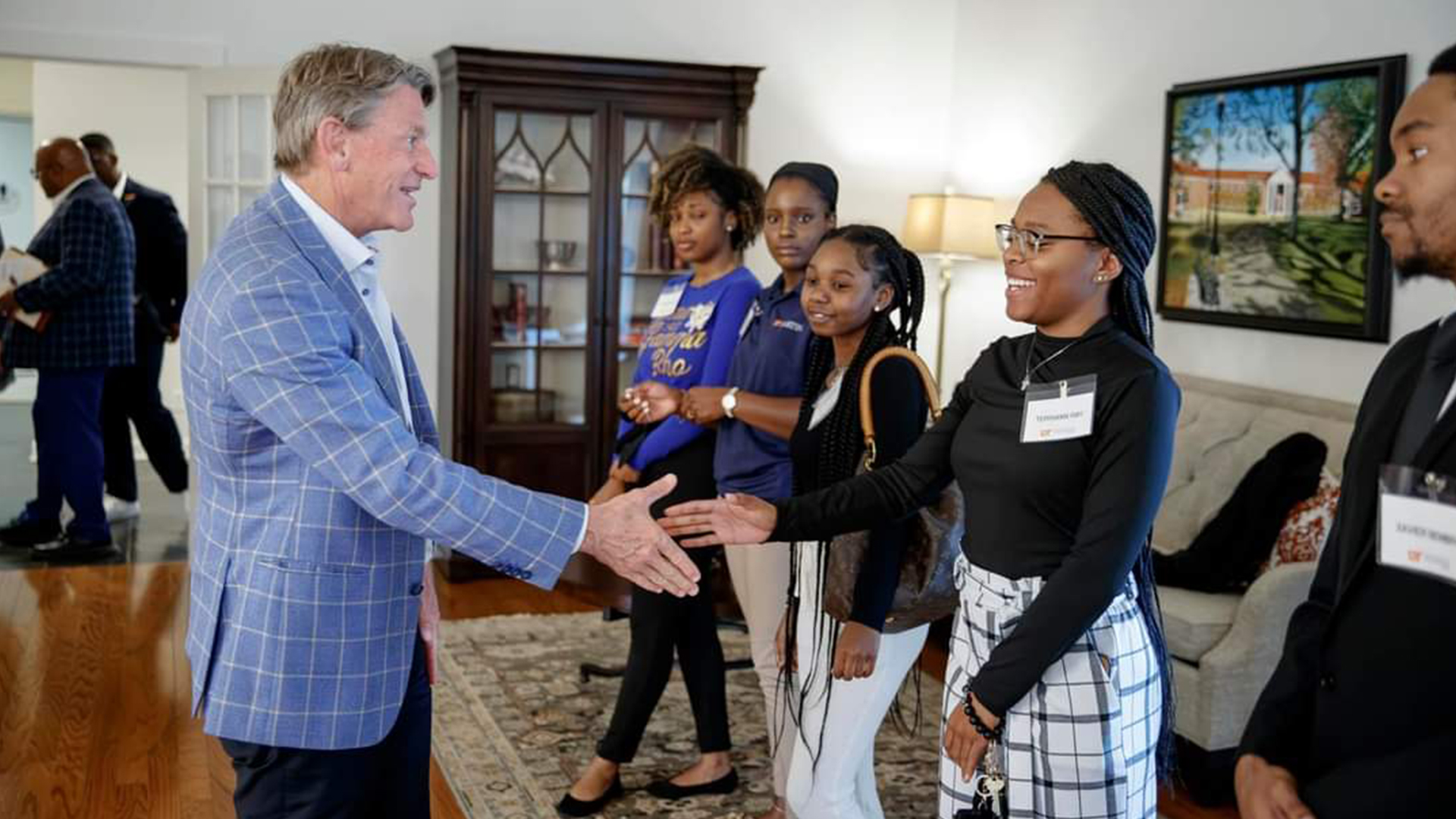 A white man in an plaid blazer shakes hands with a group of young, black students.