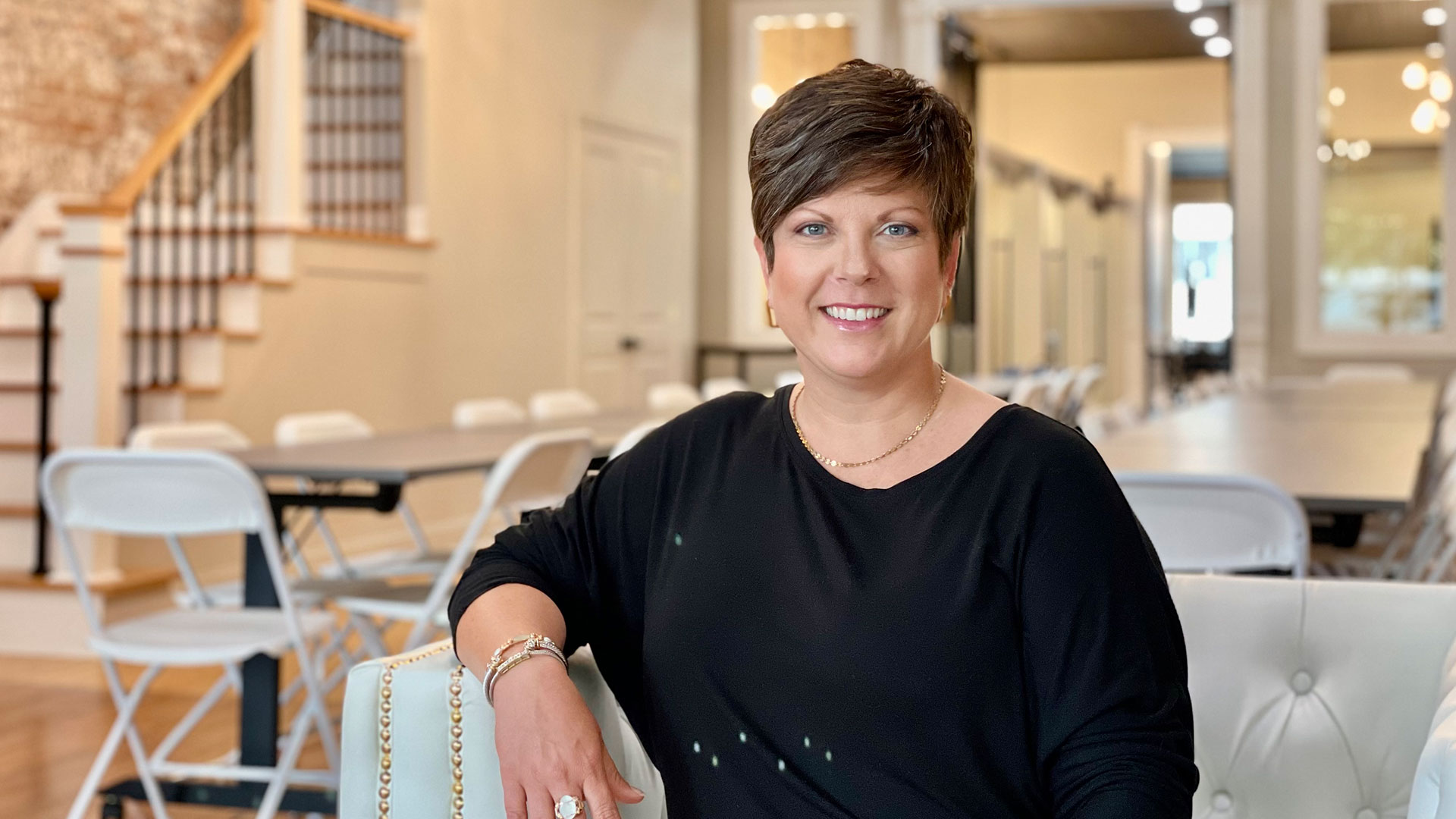 A white woman, with short brown hair and wearing a black shirt, sitting in a room with white furniture.