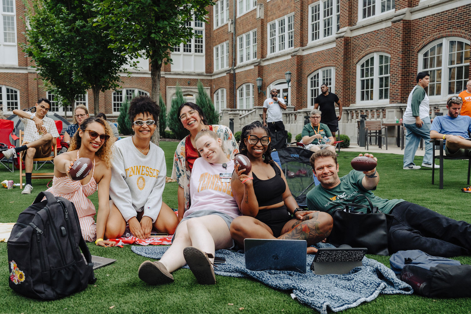 Diverse group of students and staff members lounge on a campus lawn holding small footballs and laptops.