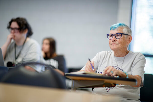 An old woman, with glasses and streaks of blue-dyed hair, sits in a classroom desk taking notes.