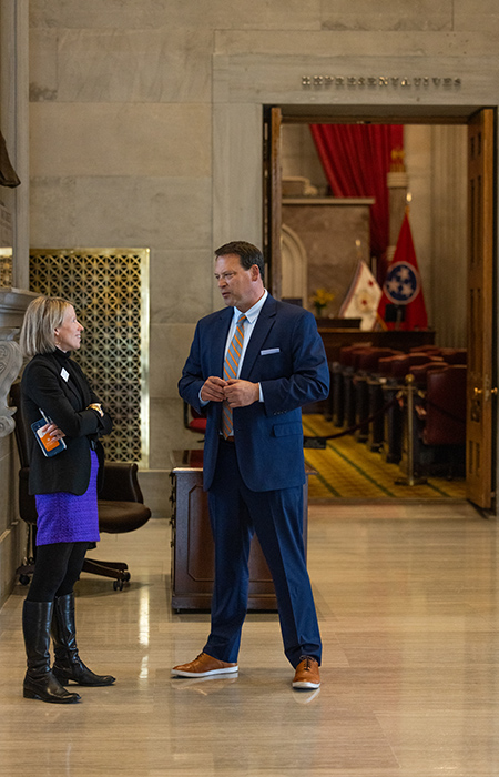 A white man in a blue suit talks with a woman wearing a black and blue outfit inside of a marble hallway.