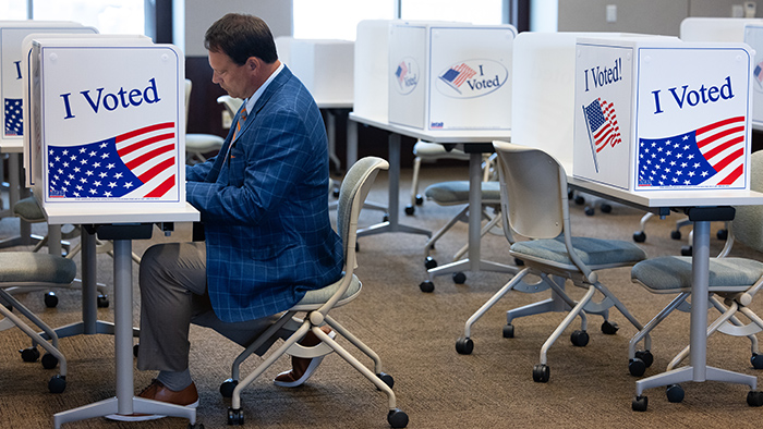 A man wearing a suit completes his voting paperwork in a room with desks and blinders labeled "I Voted."