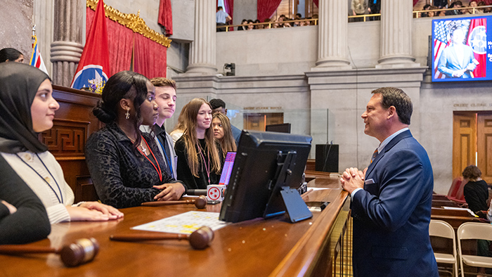 A suit-wearing man stands across from a group of students in a legislative meeting room.