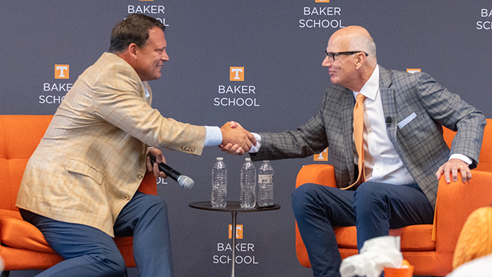 Two men wearing suits sit in orange chairs, shaking hands.