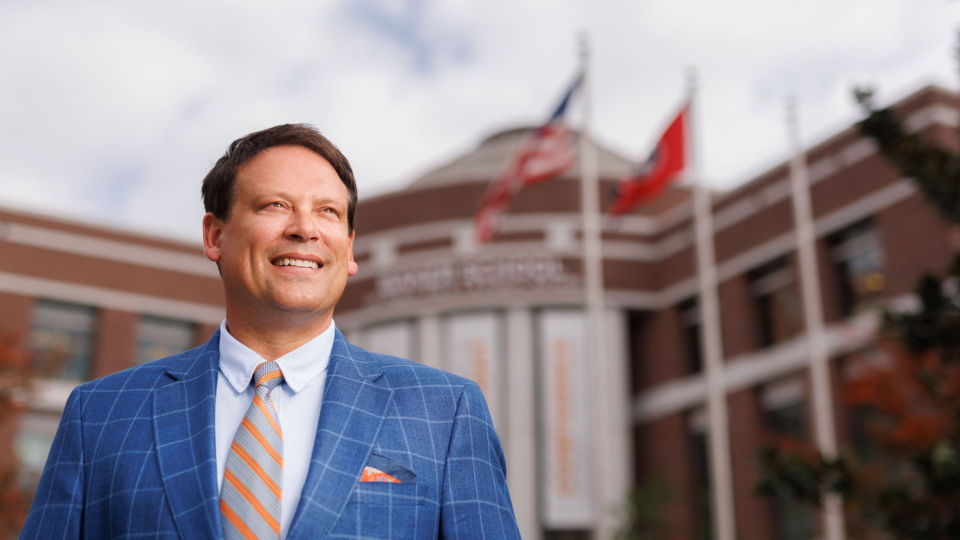 A man wearing a blue blazer and orange tie, with a building and flag blurred in the background.