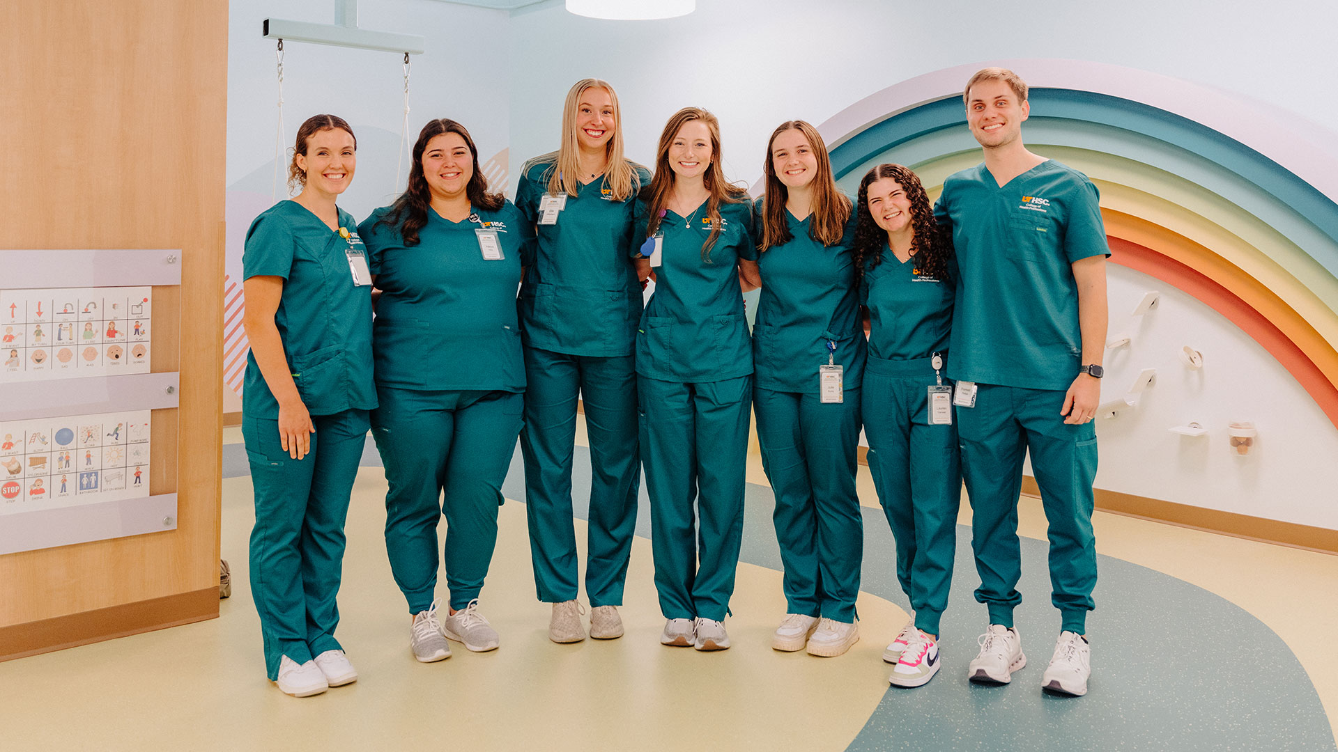 A group of six women and one man, all wearing green scrubs, standing together in a brightly decorated room.