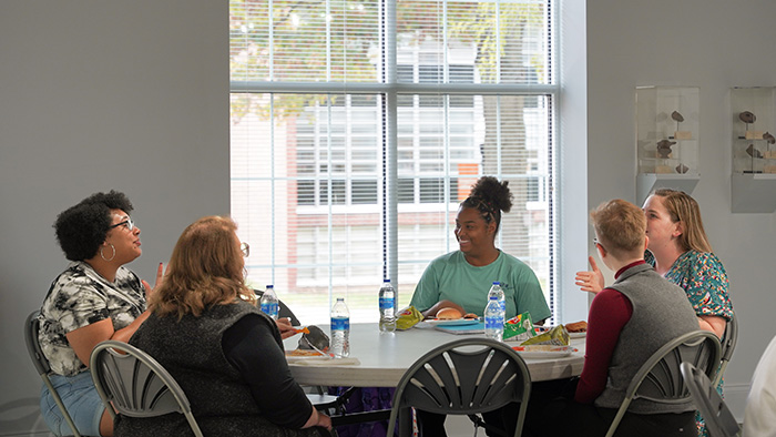 A diverse group of students sit around a table having a discussion while eating lunch.