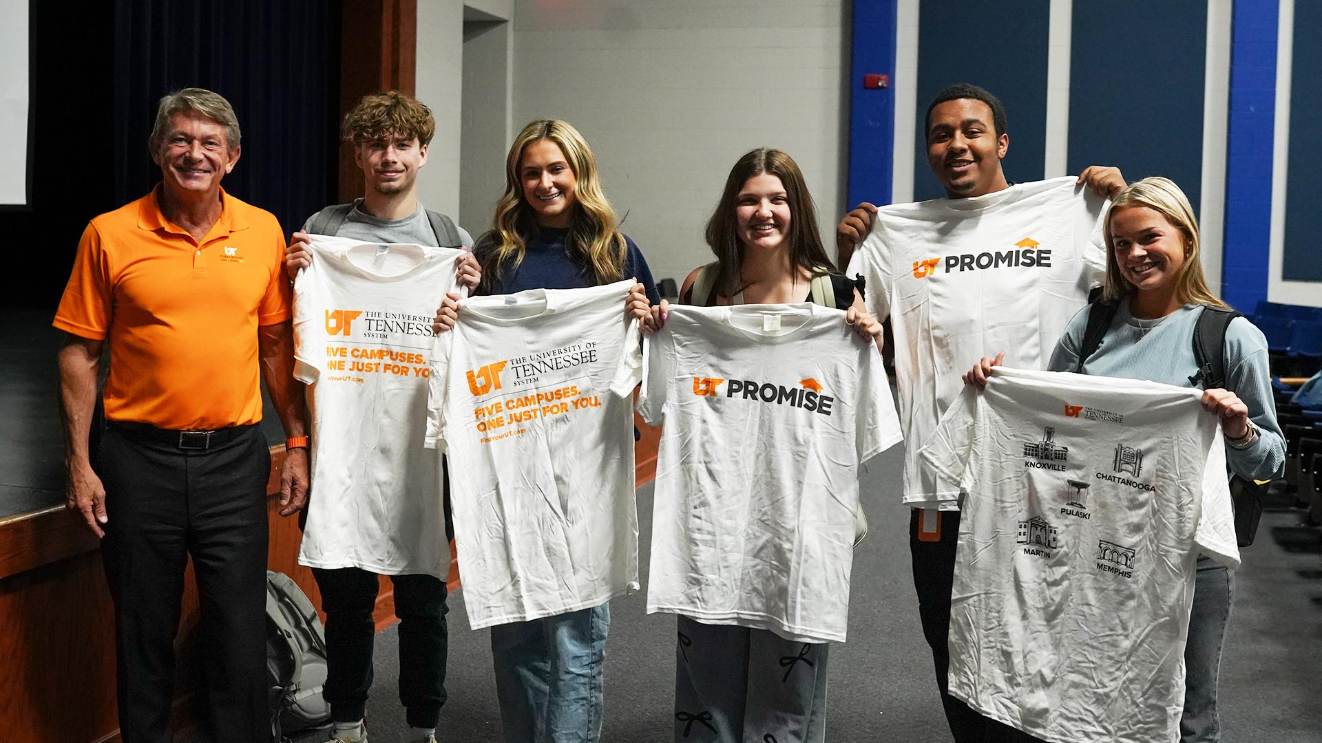 An older man, wearing an orange shirt, stands with five students holding up UT-branded shirts.