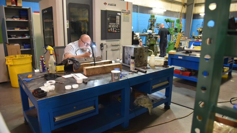 A bald man with glasses sits behind a work-table using some machinery tools.