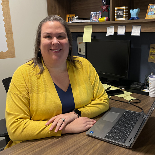 A white woman wearing a yellow blazer and blue shirt sits behind a desk, arms crossed and smiling.