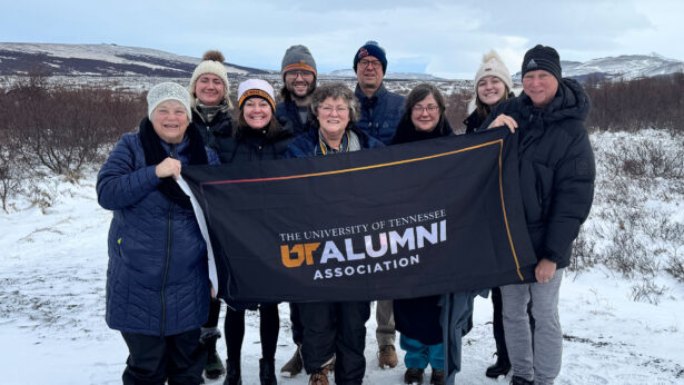 A mixed group of men and women, all wearing winter clothing, stand in a frozen landscape for a photo while holding a black flag which reads "The University of Tennessee Alumni Association."