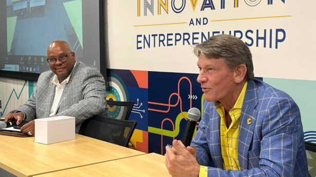 A white man, wearing a blue and yellow suit, uses a microphone while sitting next to a black man, wearing a white and gray suit.