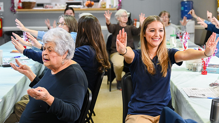 A young, blonde woman leads a group of older women through some physical therapy exercises.