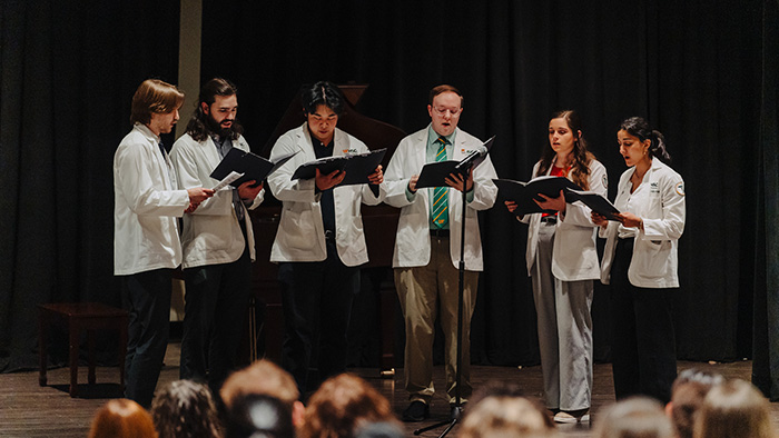 A group of six medical students, wearing lab coats, sing during a ceremony.