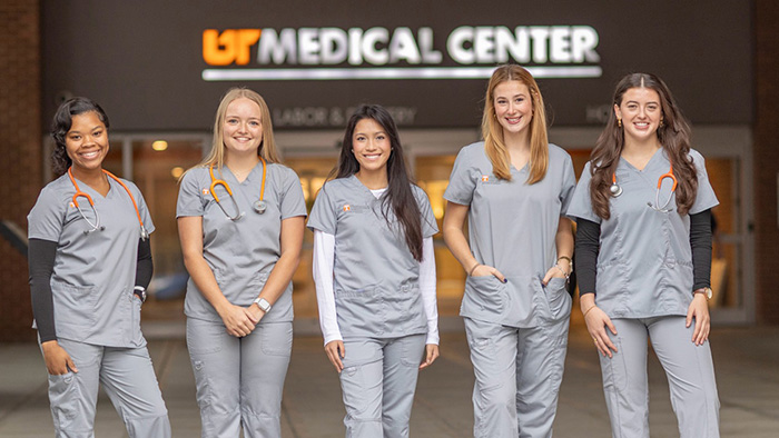 Five female students, wearing light gray scrubs, stand in-front of a sign that reads "UT Medical Center."