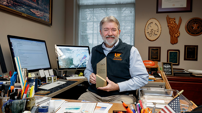 An older man with white hair and beard smiles while holding an award.