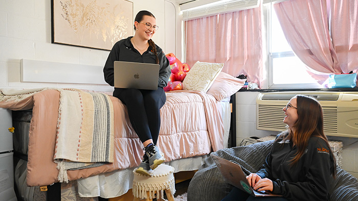 Two girls sit in their dorm rooms while using laptops.