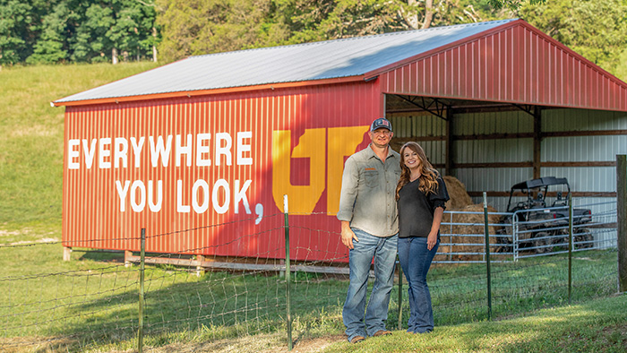 A man and woman stand in-front of a red barn, with "Everywhere You Look, UT" painted on the side.