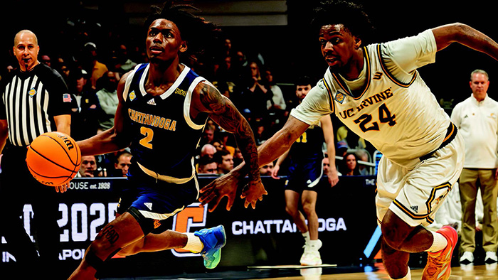 A male, black athlete in a blue jersey dribbles a basketball down court, while being chased by another black athlete in a white jersey.