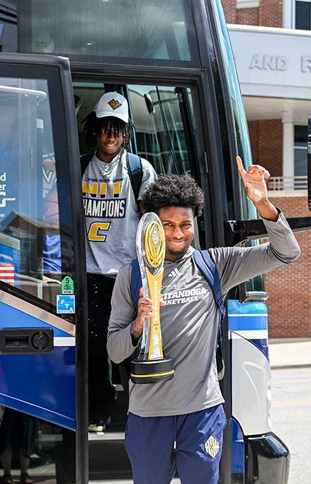 Two black athletes disembark from a tour bus, one holding a large championship trophy.