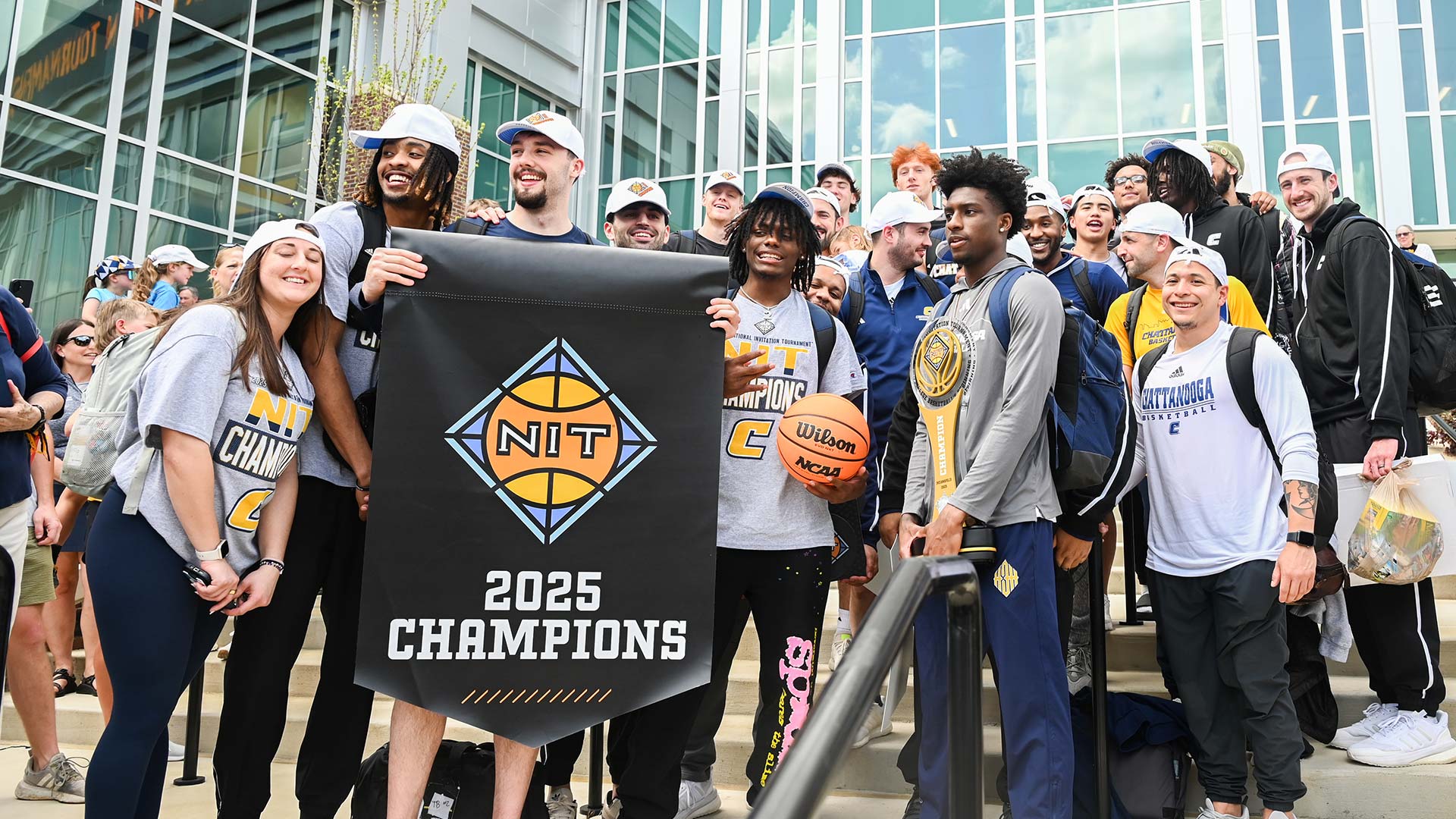 A group of college athletes standing on a stairway, holding up a banner that says "NIT 2025 Champions."