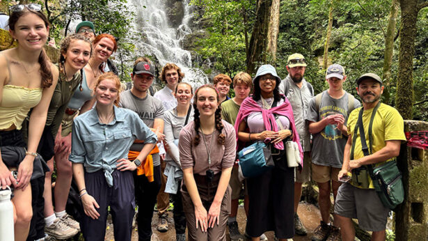 A group of students taking a photo while on a hike, with a waterfall in the background.