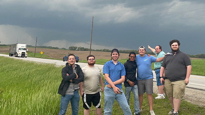A group of students take a picture on the roadside, with a tornado visible in the horizon behind them.