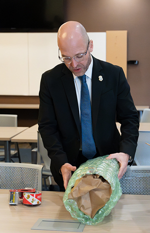 A bald man, wearing glasses and a black and white suit, wraps a package in bubble wrap in preparation for shipping.