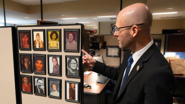 A bald man, wearing a suit and glasses, points at a collection of framed portraits of cold case victims.