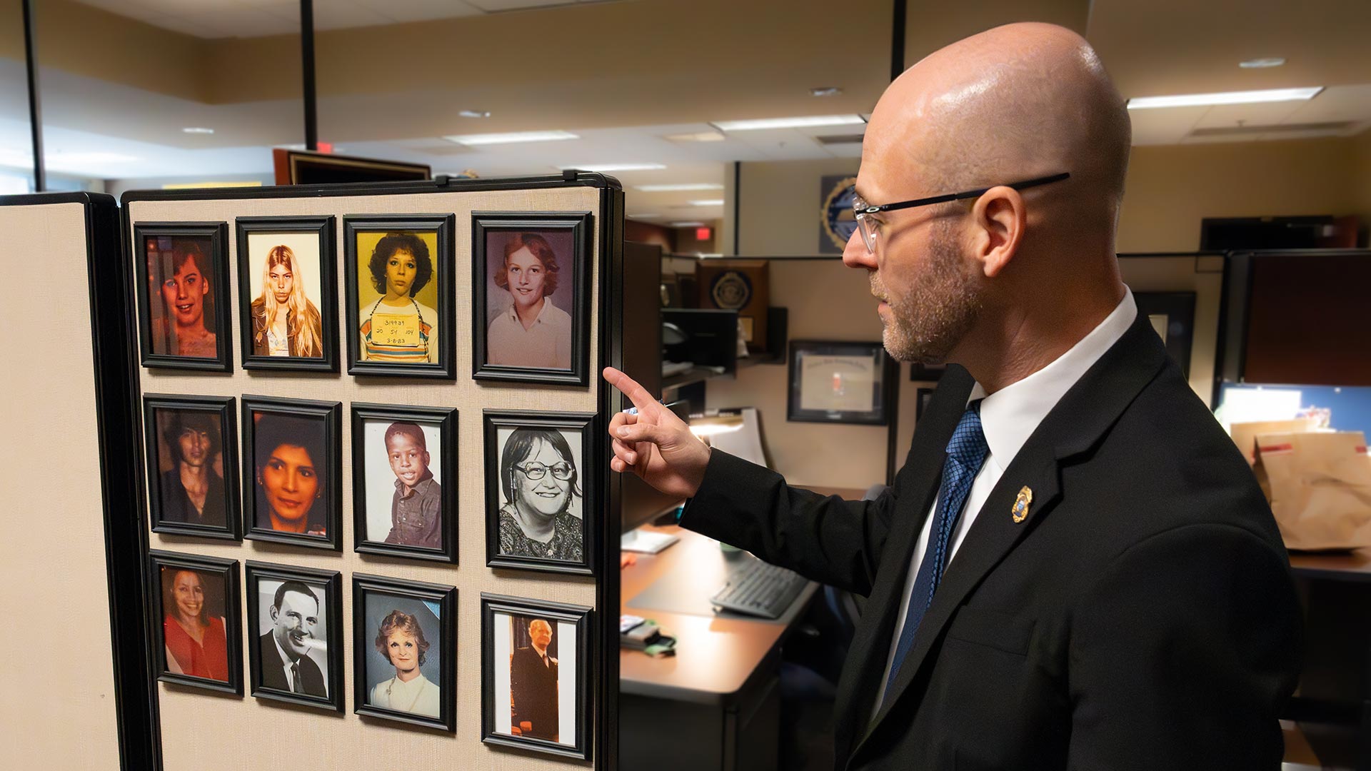A bald man, wearing a suit and glasses, points at a collection of framed portraits of cold case victims.