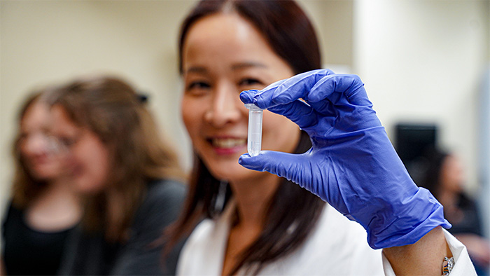 An asian woman, wearing a lab coat and blue gloves, holds up a small plastic vial.