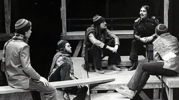 An old black and white photo of a group of young men sitting around the stage of a theatre production.