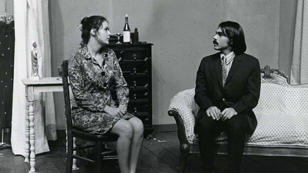 An old black and white photo of two people sitting on a couch as part of a theatre production.