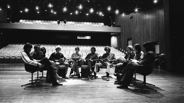 An old black and white photo of a group of actors sitting around the stage during a script reading of a theatre production.