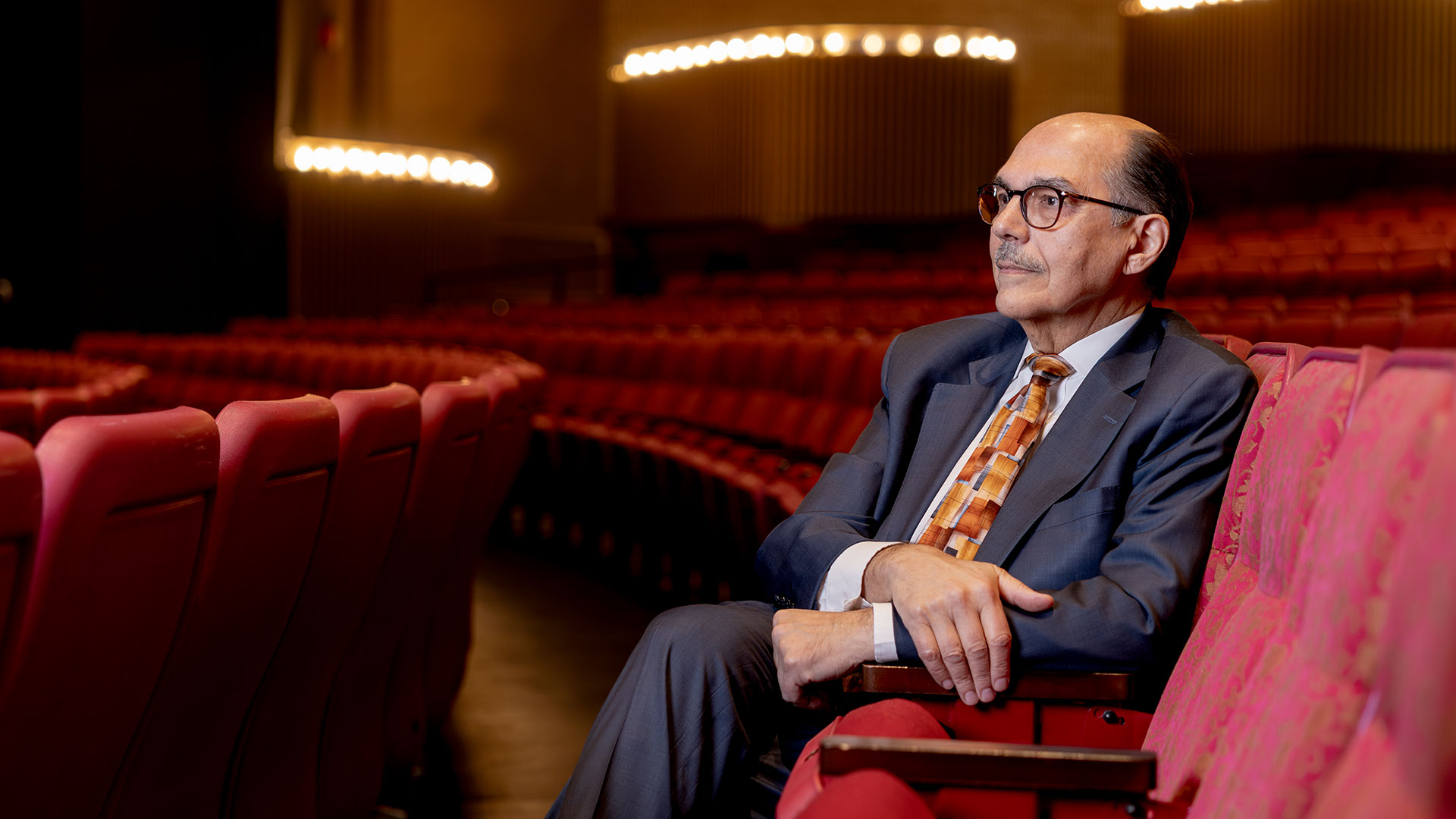 A man, wearing glasses and a gray suit, sits alone in a theatre filled with rows of red seats.
