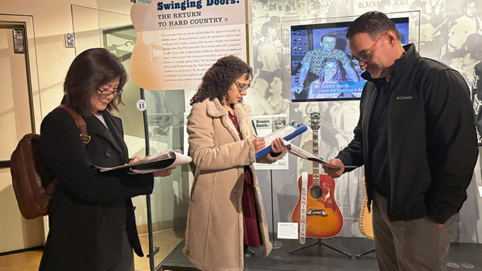 A man and two women all stand while reading printed materials in front of a music museum display.