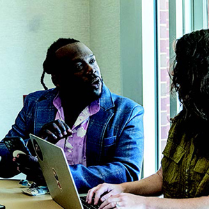 A black man, wearing a blue blazer, sits at a table and talks with a woman who is using a laptop.