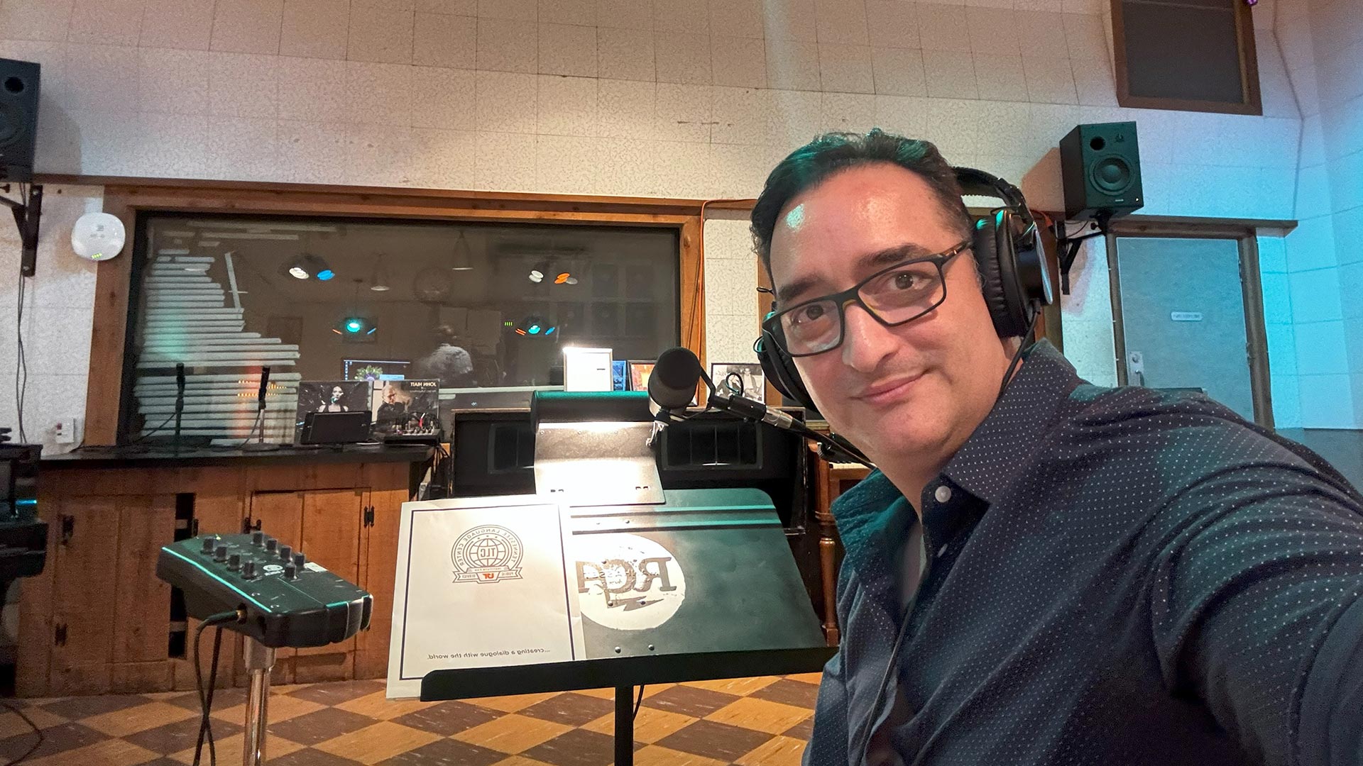A man wearing glasses and a blue shirt sits inside a recording studio with headphones on.