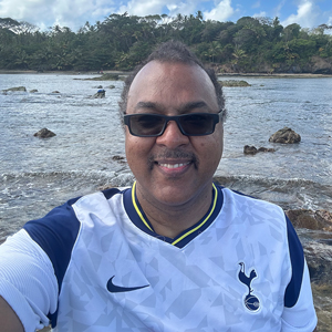 A man wearing sunglasses stands on a beach, with the ocean and an island behind him.