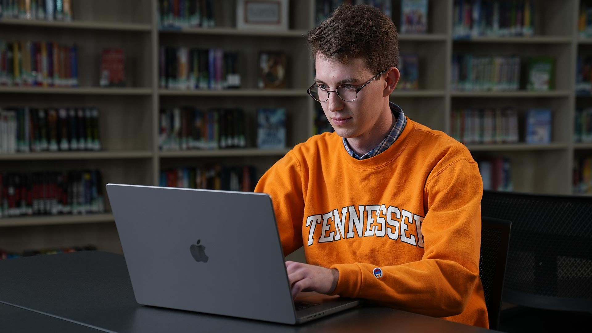 A man with glasses, wearing an orange 'Tennessee' sweatshirt, sits working on a laptop.